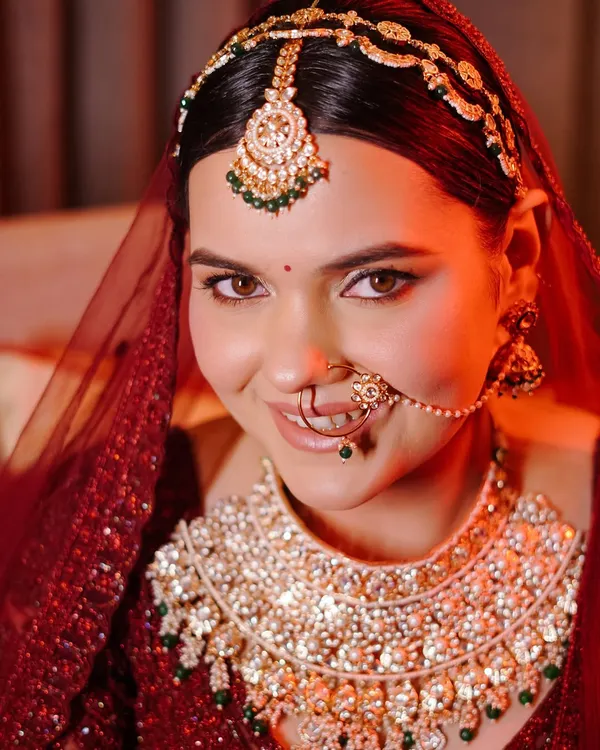 A stunning close-up of the bride, her face illuminated by a warm red light. This portrait beautifully captures her flawless makeup, intricate nose ring, and the heavy diamond and emerald necklace.