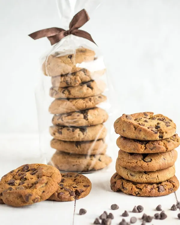 A stack of our classic Chocolate Chip Cookies, with a bag of them ready for gifting in the background. These cookies are soft, chewy, and packed with chocolate chips, a timeless favourite.