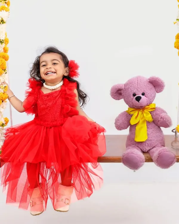 A picture of pure joy. This happy toddler is dressed in a bright red dress, sitting on a flower-adorned swing with her teddy bear companion for a fun and whimsical portrait.