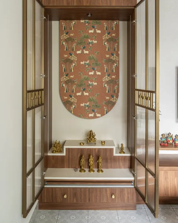 This elegant puja unit features a beautiful wallpaper backdrop with a traditional Indian motif. The unit itself has stepped platforms for idols and is enclosed by doors with fluted glass and metal accents.