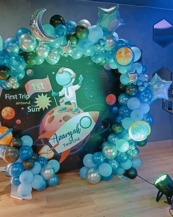 A "First Trip Around the Sun" space theme for a first birthday. The circular backdrop shows an astronaut on a rocket, surrounded by a balloon arch designed to look like the solar system.