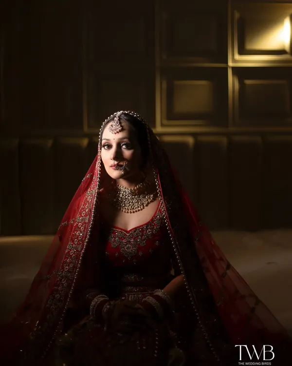 A stunning bridal portrait in full color. The bride, dressed in a traditional red lehenga, looks directly at the camera with a calm and graceful expression, her beauty illuminated by soft, warm light.