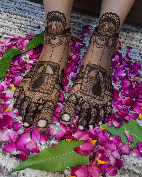 A stunning bridal leg design featuring a peacock portrait within a frame, surrounded by intricate patterns and a lotus anklet band. The design is beautifully presented on a bed of flower petals.