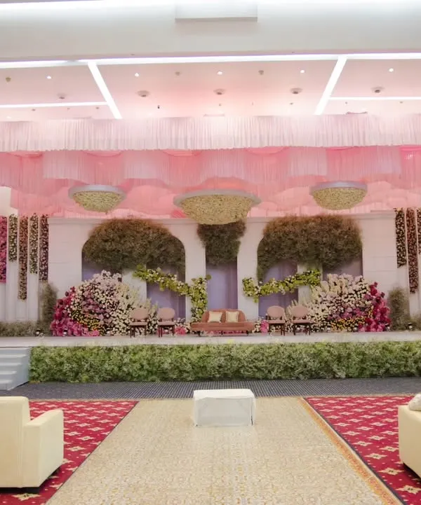 A wide view of a reception stage decorated in soft pinks and lush greenery. The design features elegant draping and floral arrangements that create a romantic and sophisticated backdrop for the main event.