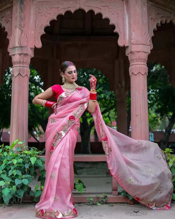 A full-length portrait of a woman in a flowing pink organza saree, standing gracefully in a heritage-style gazebo. The shot captures the elegance of the saree and the beauty of the outdoor location.
