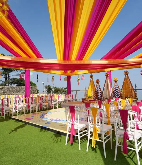 An expansive view of a stunning rooftop Haldi ceremony. We used vibrant yellow and pink drapes to create a festive canopy against the blue sky, with a beautifully decorated stage and seating for all the guests.