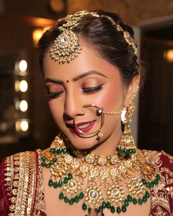 A classic bridal portrait showcasing a flawless HD base, warm smokey eyes, and a rich red lip. The look is completed with traditional gold and green kundan jewelry, perfect for a timeless wedding ceremony.