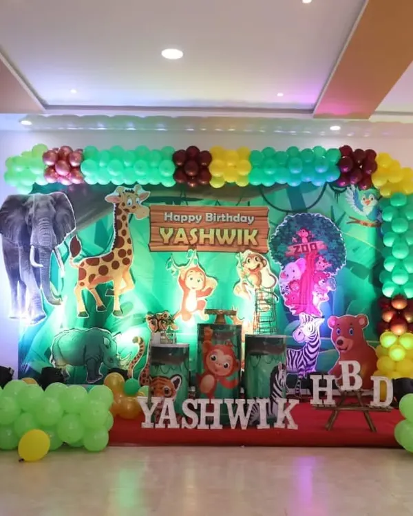 A fun jungle safari theme for Yashwik's birthday. This stage decoration includes a vibrant green backdrop, cutouts of various jungle animals like a giraffe and elephant, and a matching green balloon frame.