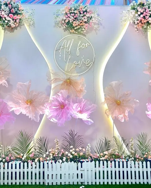 This modern and artistic photo booth features a backdrop with large, handcrafted paper flowers in shades of pink and peach. A custom neon sign with the phrase "All for Love" and soft backlighting creates a romantic and stylish spot for photos.