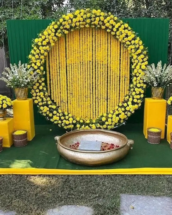 A classic Haldi backdrop for outdoor functions. This setup features a large circular frame filled with strings of yellow marigolds, a green panel background, and a traditional brass urli in the center for the ceremony.