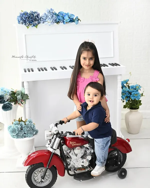 A fun and musical sibling portrait. The baby boy takes a ride on a toy motorcycle while his big sister stands by the piano, creating a playful and charming scene in our all-white studio setup.
