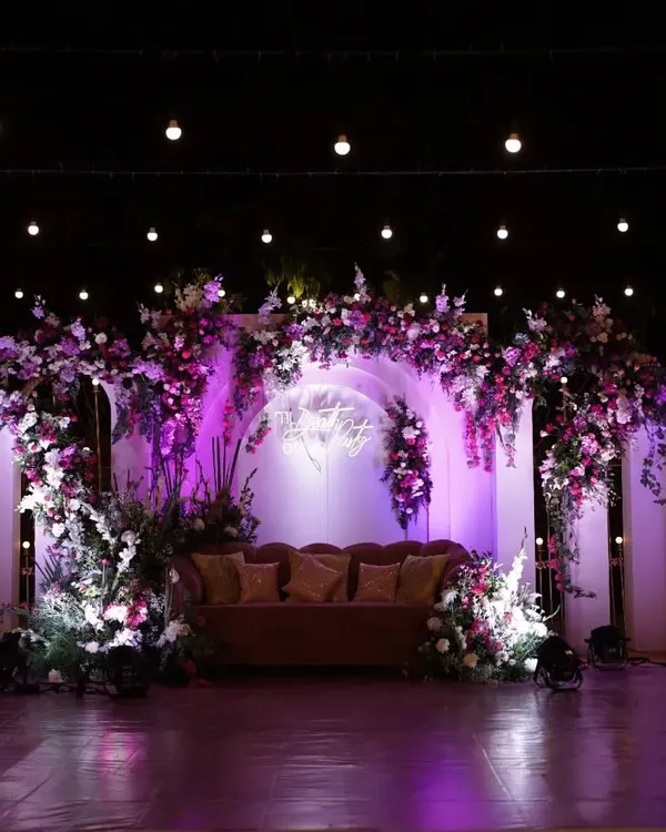 A dramatic and romantic reception stage featuring a lush floral arch in shades of purple and white. The custom neon sign and strategic uplighting create a vibrant, modern, and photo-ready backdrop.