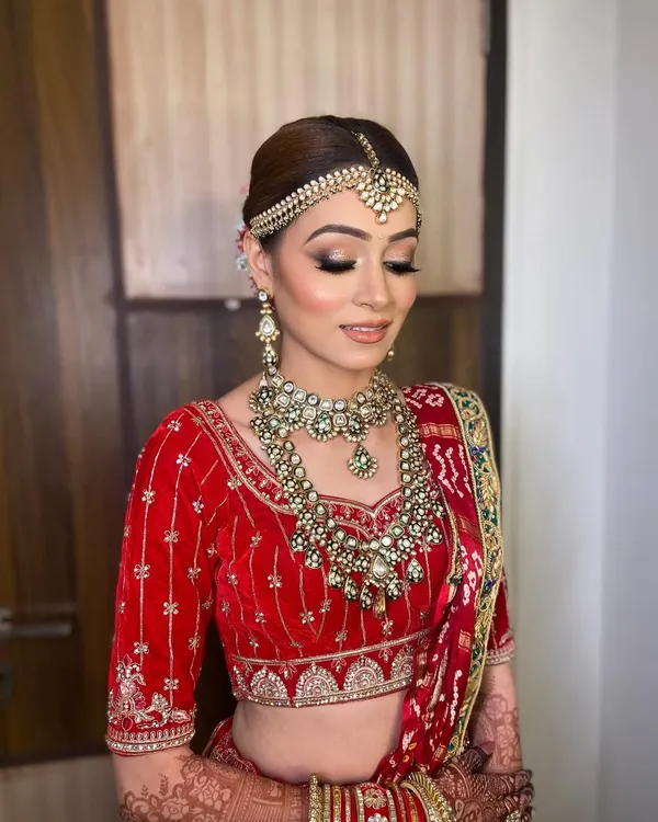 A classic red bridal look for Bhavika, featuring a flawless base and defined eyes to complement her traditional velvet lehenga and heavy jewelry.