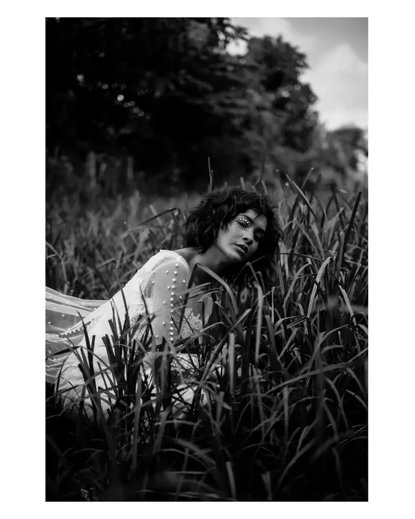 A dramatic black and white portrait in a field of tall grass. The high contrast and the model's pose create a moody, cinematic, and slightly mysterious feel.
