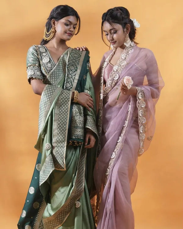 A beautifully composed shot of two models, showcasing a sage green silk saree and a lavender organza saree. This image is perfect for a lookbook cover, demonstrating how different pieces from a collection can be styled together.