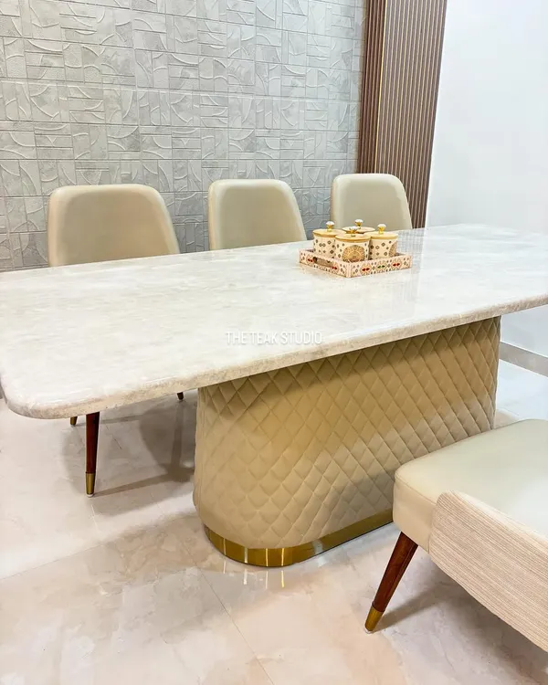 This modern dining set features a stunning Italian marble tabletop with softly rounded corners. The custom base is upholstered in a neutral quilted fabric with a brass-finished metal trim, perfectly complemented by elegant chairs with tapered wooden legs.