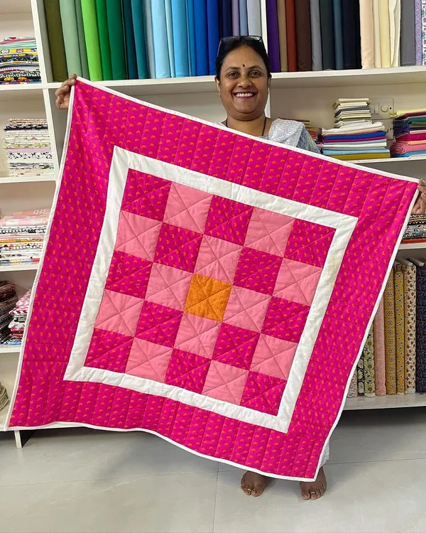 A happy student from our Beginner's Quilting Class holds up her first project, a vibrant pink and orange checkerboard quilt.