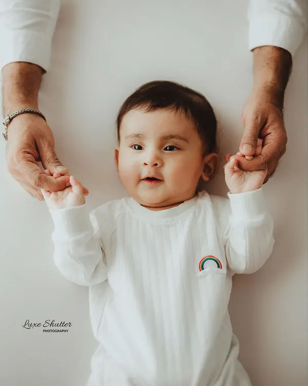 A sweet five-month milestone photo, with the baby lying down and holding his parent's hands. His direct gaze and cute expression are priceless.