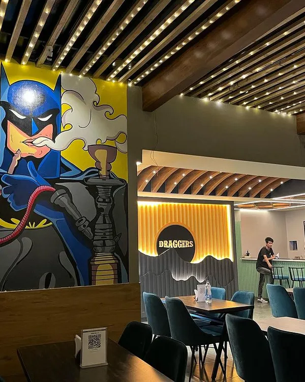 Welcome to the all new Draggers Cafe. This wide view showcases our revamped interior, featuring modern wooden ceiling panels, comfortable teal seating, and our iconic Batman mural, making it the perfect backdrop for your hangout.