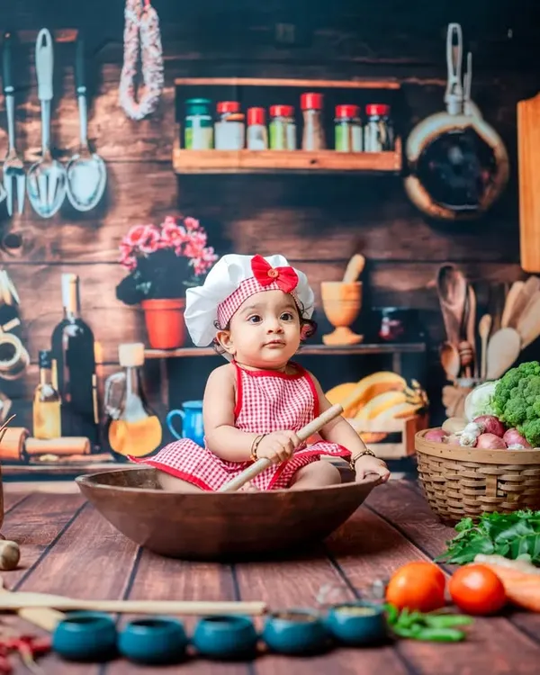A curious little chef inspects her ingredients. The 'Little Chef' theme is perfect for sitter sessions, allowing babies to safely interact with various props like wooden spoons and fresh vegetables in a charming kitchen setup.