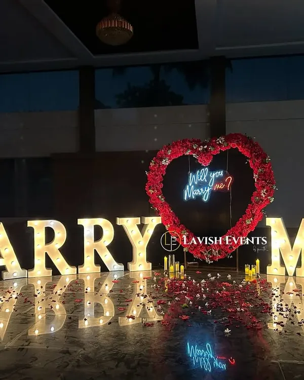 The perfect proposal setup. Giant illuminated "MARRY ME" letters, a heart-shaped arch made of red roses, and a neon "Will you Marry me?" sign create a breathtaking scene.