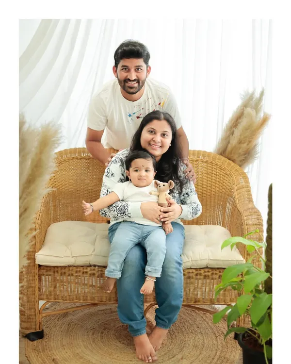 A warm and relaxed family portrait in a studio with natural-looking props. The parents and their baby are dressed in casual whites and blues, creating a cohesive and timeless look.