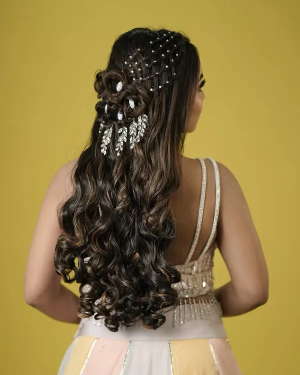 An intricate and glamorous half-updo with cascading curls, detailed with a pearl net and leaf-like crystal accessories. A true showstopper for a grand reception.