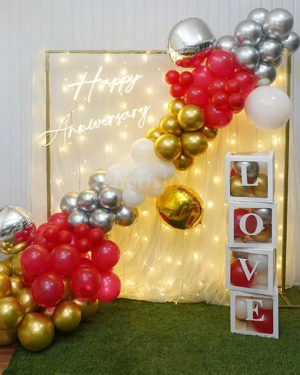 A popular choice for home anniversaries, this decor combines a trendy 'Happy Anniversary' neon sign with a backdrop of warm fairy lights. The diagonal balloon garland in red, gold, and silver adds a splash of color and fun.