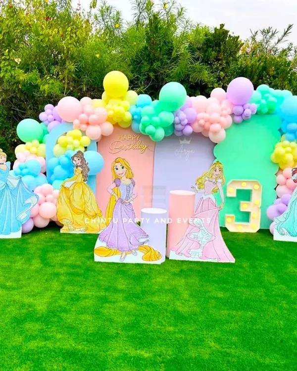 A princess-themed birthday party featuring character cutouts of Cinderella, Belle, and others against a pastel backdrop. The setup includes a balloon arch and a light-up marquee number for the birthday girl.