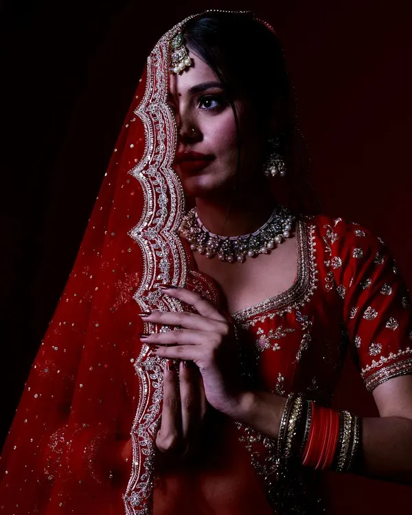 A classic bridal pose with the dupatta, creating a sense of mystery and elegance. This studio portrait uses focused lighting to draw attention to the bride's expressive eyes and the detailed embroidery of her red veil.