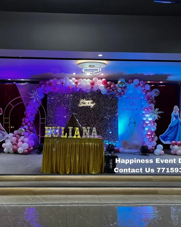 A grand princess theme for a 5th birthday in a banquet hall. This premium setup features a full stage with a silver sequin wall, a balloon arch in shades of pink and chrome, and life-sized cutouts of popular princesses.
