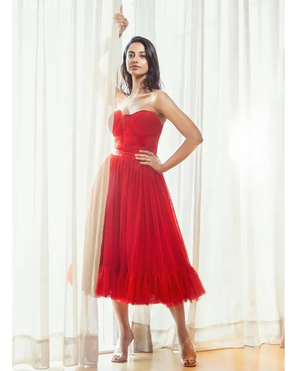A striking full-length shot of a red tulle gown. The styling is clean and modern, using natural light from a window to create a soft yet dramatic effect.