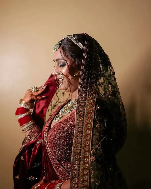 A side profile of a bride in a deep maroon velvet lehenga, showcasing the intricate embroidery and the complete look with a sheer dupatta. This is a classic choice for a winter wedding theme.