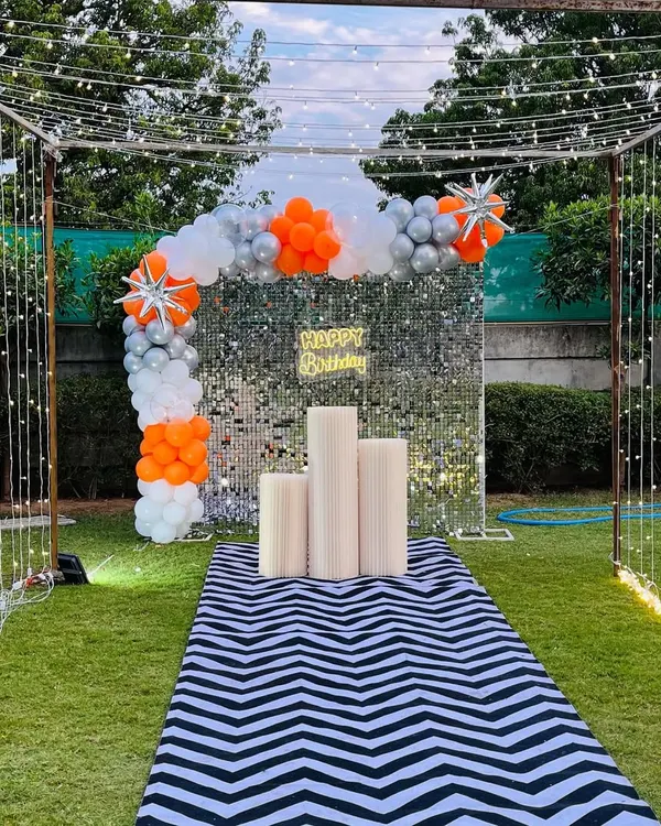 A stunning garden party entrance with a walkway leading to a silver shimmer wall, framed by fairy lights and an orange and silver balloon arch.