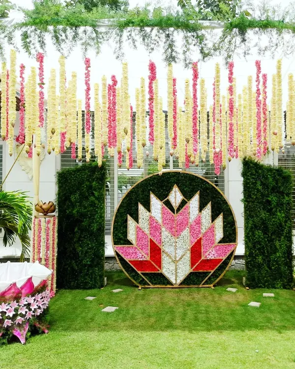 A modern and artistic naming ceremony setup in a garden. The centerpiece is a large circular frame with a geometric lotus pattern made of flower petals, flanked by green walls and hanging floral strings.