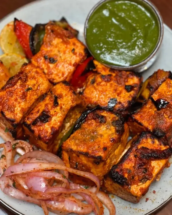 My Paneer Tikka features large, soft cubes of paneer marinated in yogurt and spices, then grilled with bell peppers and onions. It's served with a side of spicy green chutney and pickled onions for a complete appetizer experience.