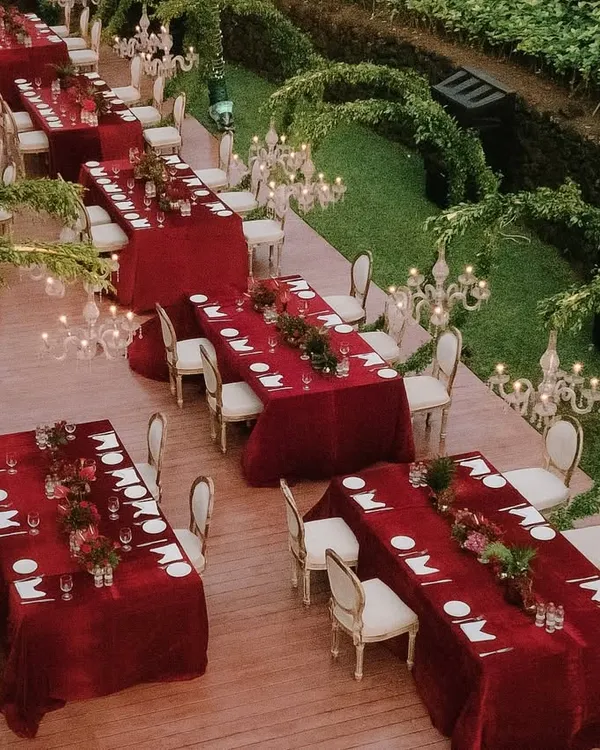 An aerial view of a spectacular engagement dinner setup in Goa. The rich red tables are arranged on a raised platform, surrounded by lush greenery and illuminated by elegant candelabras, creating a truly royal ambiance.
