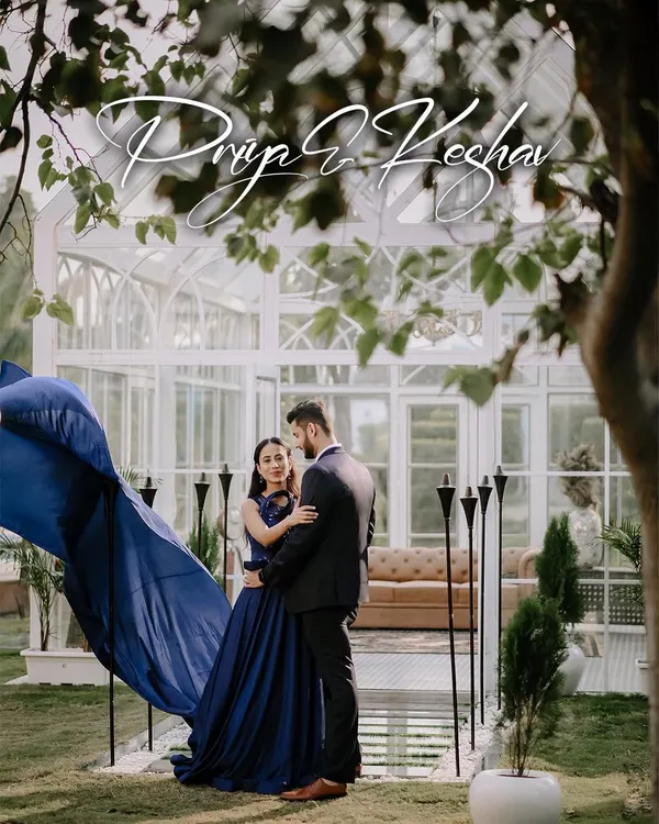 A dramatic pre-wedding portrait featuring a long, flowing blue gown. The movement of the fabric against the elegant white greenhouse creates a stunning, high-fashion look.