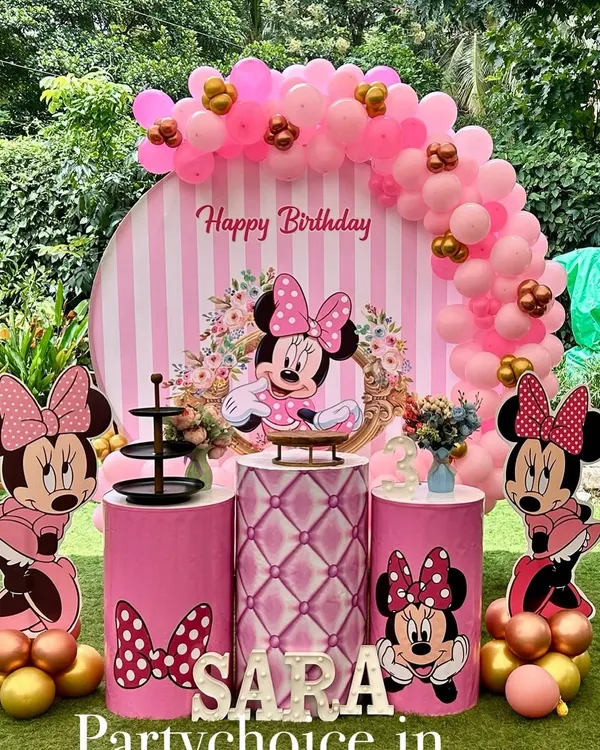 A classic Minnie Mouse birthday party setup in an outdoor garden setting. This decoration features a pink and white striped circular backdrop, a pink balloon arch with gold accents, and multiple Minnie cutouts.