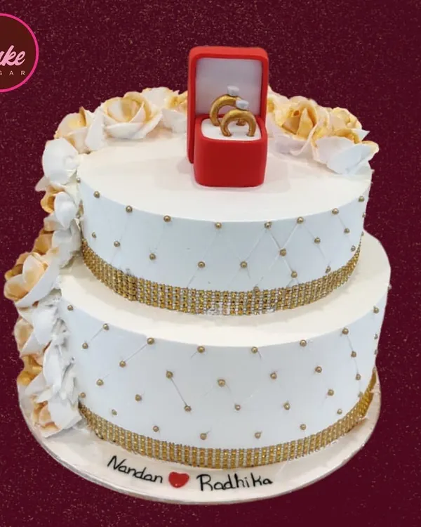 A beautiful two-tiered engagement cake in white and gold. The design features a quilted pattern, sparkling bands, and is topped with an edible ring box, symbolizing the special commitment.
