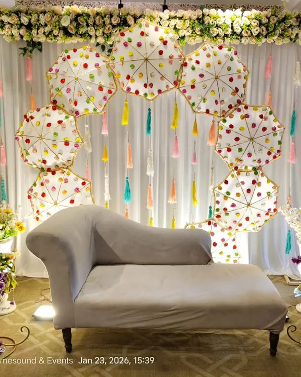 A unique Mehndi backdrop featuring an arch of decorative white umbrellas and colorful tassels. The floral border at the top and side arrangements complete this fun, mela-style setup.