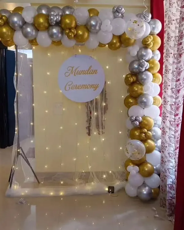An elegant and modern Mundan ceremony backdrop. This setup features a white and gold balloon frame, a personalized sign, and a delicate curtain of fairy lights.