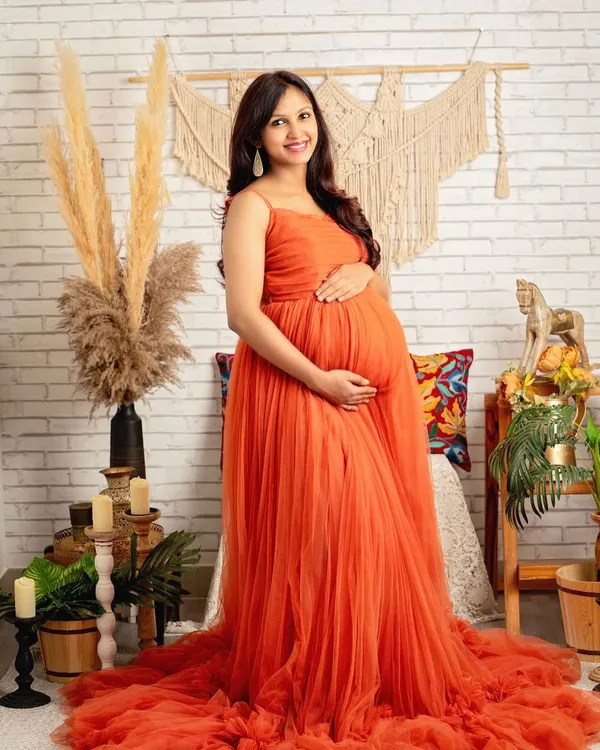 The vibrant orange gown against the rustic brick and macrame backdrop creates a stunning, warm portrait. Her smile says it all.