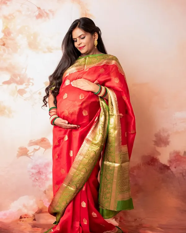 Embracing tradition and elegance, this studio maternity portrait showcases a stunning red and green silk saree. The soft, painted backdrop adds a touch of artistry to this classic look.