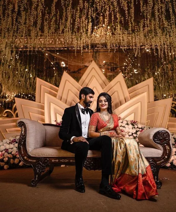 A formally dressed couple is elegantly posed on a luxurious chaise lounge against a grand, lotus-themed stage at their wedding reception. We specialize in capturing the scale and design of large-scale wedding events.