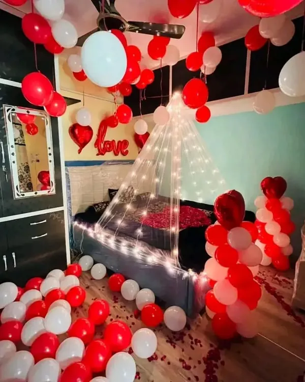 A classic red and white themed room decoration. The space is filled with balloons on the floor and ceiling, with a fairy light canopy over the bed and 'Love' foil balloons to complete the romantic surprise.