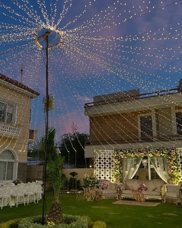 A stunning outdoor setup for an anniversary celebration at dusk. The canopy of lights, elegant seating, and floral stage create a truly magical and romantic atmosphere.