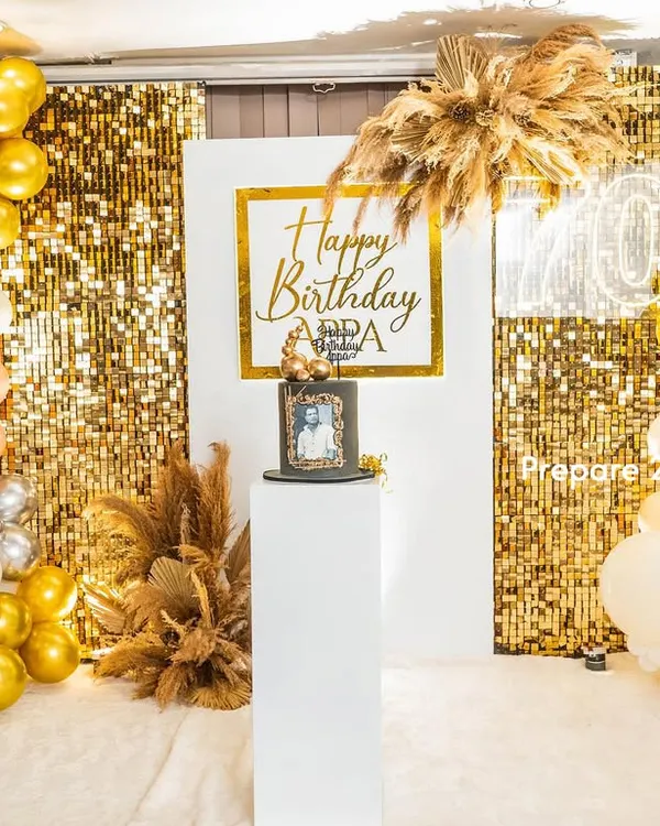 A chic and modern backdrop for a 70th birthday celebration. This setup features a gold shimmer wall, a white central panel with custom calligraphy, and organic balloon arches in gold, silver, peach, and white, accented with dried pampas grass.