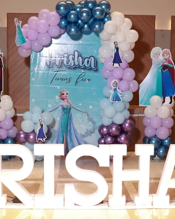 A magical Frozen-themed party for Prisha's fifth birthday. The backdrop features Elsa and Anna, framed by a balloon arch in icy shades of blue, purple, and white, with large marquee letters in the front.