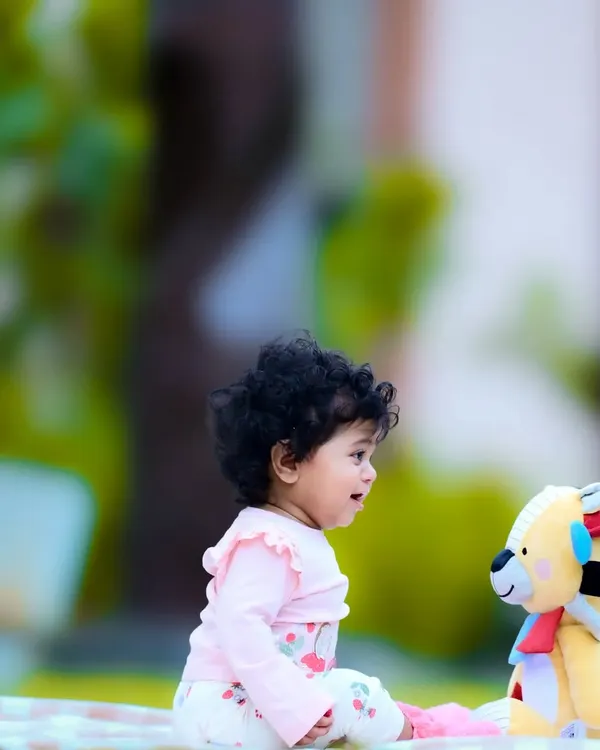 A baby sitting on a picnic blanket outdoors, having a sweet conversation with a teddy bear.